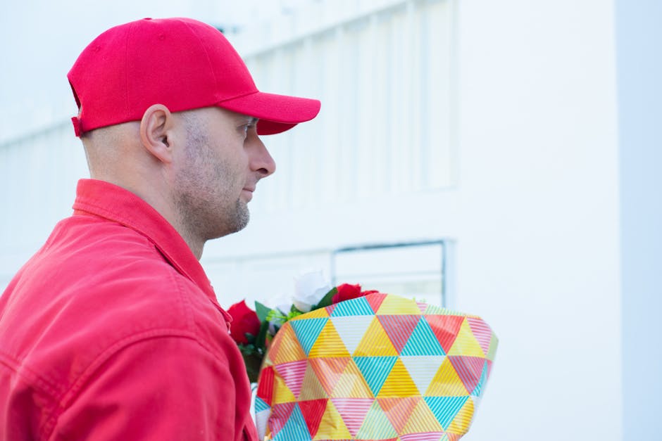 A man wearing a red cap and red jacket holds a colorful floral gift wrapped in patterned paper with vibrant yellow, blue, and red triangles. The bouquet contains mixed flowers, including red and white