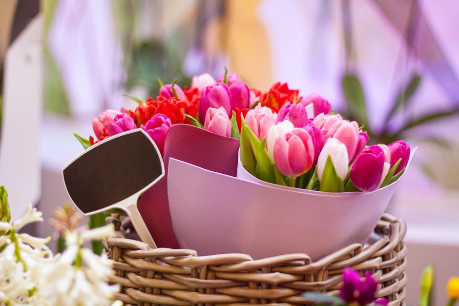 A close-up of a bouquet arrangement featuring pink and white tulips with green stems and leaves, wrapped in pastel pink and purple paper, placed inside a woven wicker basket. The bouquet is positioned