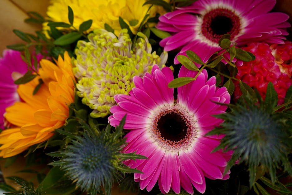 A vibrant floral bouquet arranged with a variety of fresh flowers, including large pink gerbera daisies with black centers, bright yellow chrysanthemums, green and white chrysanthemums, and blue thist
