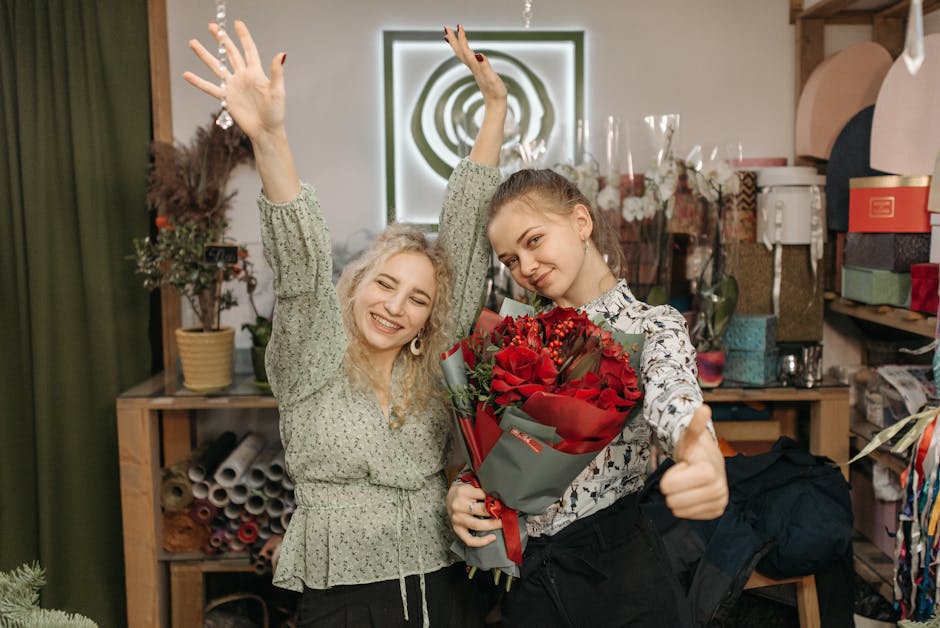 Two young women indoors in a floral workshop or flower shop environment, celebrating with a bouquet of fresh red roses wrapped in grey and red decorative paper. The woman on the right, holding the bou