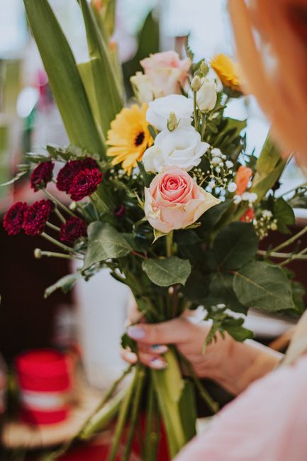 A floral bouquet featuring a variety of fresh flowers including soft pink roses, deep red carnations, white lisianthus, and bright yellow daisies, arranged with green foliage and stems. The bouquet is